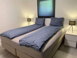 A double bed with a grey blanket, two bedside lamps, and a window with blinds.