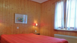 A room with wooden panel walls, a bed with a red bedspread, a nightstand with a lamp, a framed picture on the wall, and a window with curtains. - Lill Britta 2, mittlere Wohunug Appartement/Fewo, Dusche und Bad, WC, Komfort by Interhome (Arosa)
