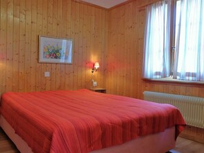 A room with wooden panel walls, a bed with a red bedspread, a nightstand with a lamp, a framed picture on the wall, and a window with curtains. - Lill Britta 2, mittlere Wohunug Appartement/Fewo, Dusche und Bad, WC, Komfort by Interhome (Arosa)