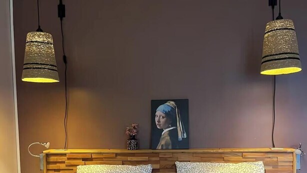 Ett sovrum med en sänggavel i trä, en säng med grönt och vitt sängklädnad, två hänglampor och en inramad tavla på väggen.