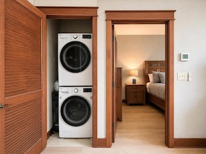 A laundry room with two stacked washing machines and a bedroom with a bed, nightstand, and lamp. - Unwind at Riverstone Loft (Coeur d'Alene)