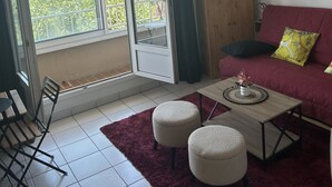 A living room with a red sofa, a coffee table, two ottomans, a wooden cabinet, and a view of greenery through large windows. - Bright, air-conditioned studio with views of the sea and mountains, 5 minutes from the harbor and downtown. (Port-Vendres)
