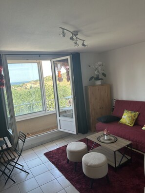 Sala de estar com sofá vermelho, mesa de centro, dois banquinhos, armário de madeira e vista para o verde através de grandes janelas.