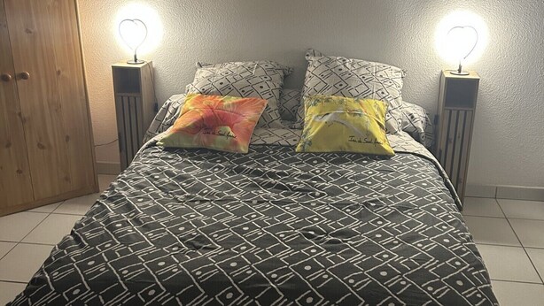 A bed with a patterned bedspread, two pillows, two bedside lamps, and a wooden wardrobe. - Bright, air-conditioned studio with views of the sea and mountains, 5 minutes from the harbor and downtown. (Port-Vendres)