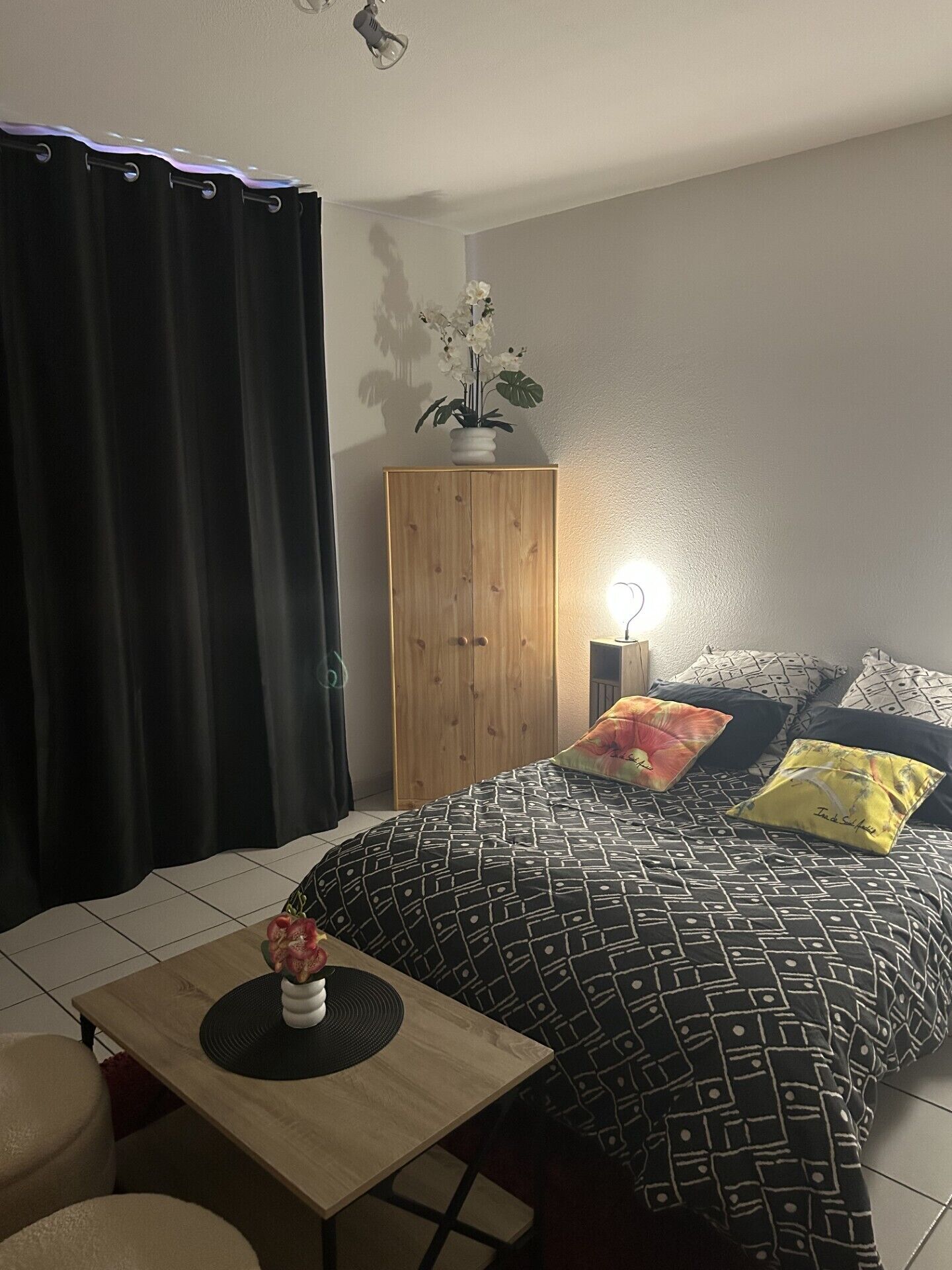 A bedroom with a bed, a wooden wardrobe, a bedside lamp, and a small table with a vase.