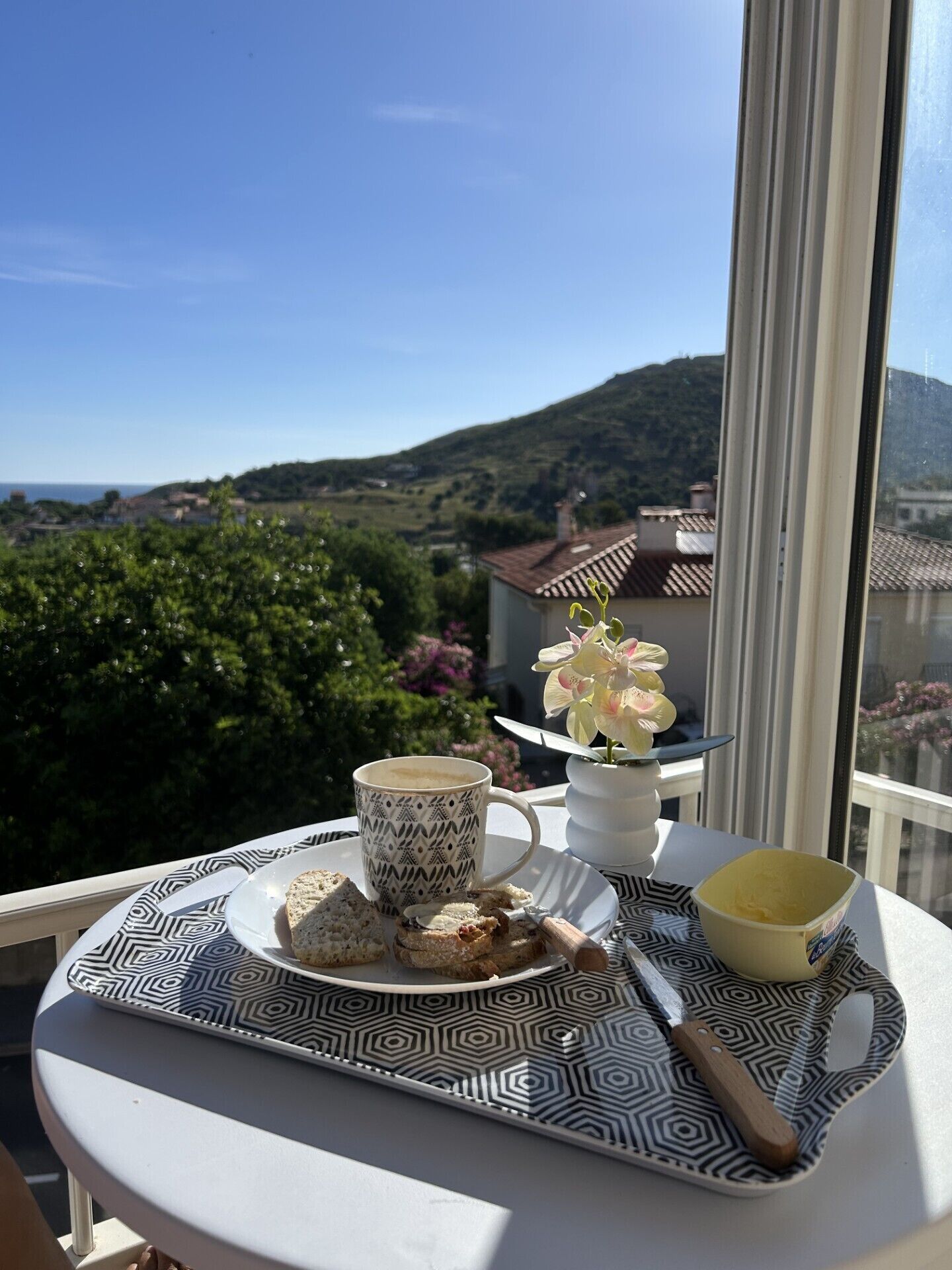 A tray with a cup, pastries, and a flower on a balcony overlooking a scenic view.