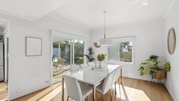 A modern dining area with a white table and chairs, a pendant light, and a view of the outdoors through large windows.
