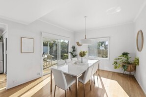 Ein moderner Essbereich mit einem weißen Tisch und Stühlen, einer Pendelleuchte und Blick ins Freie durch große Fenster.