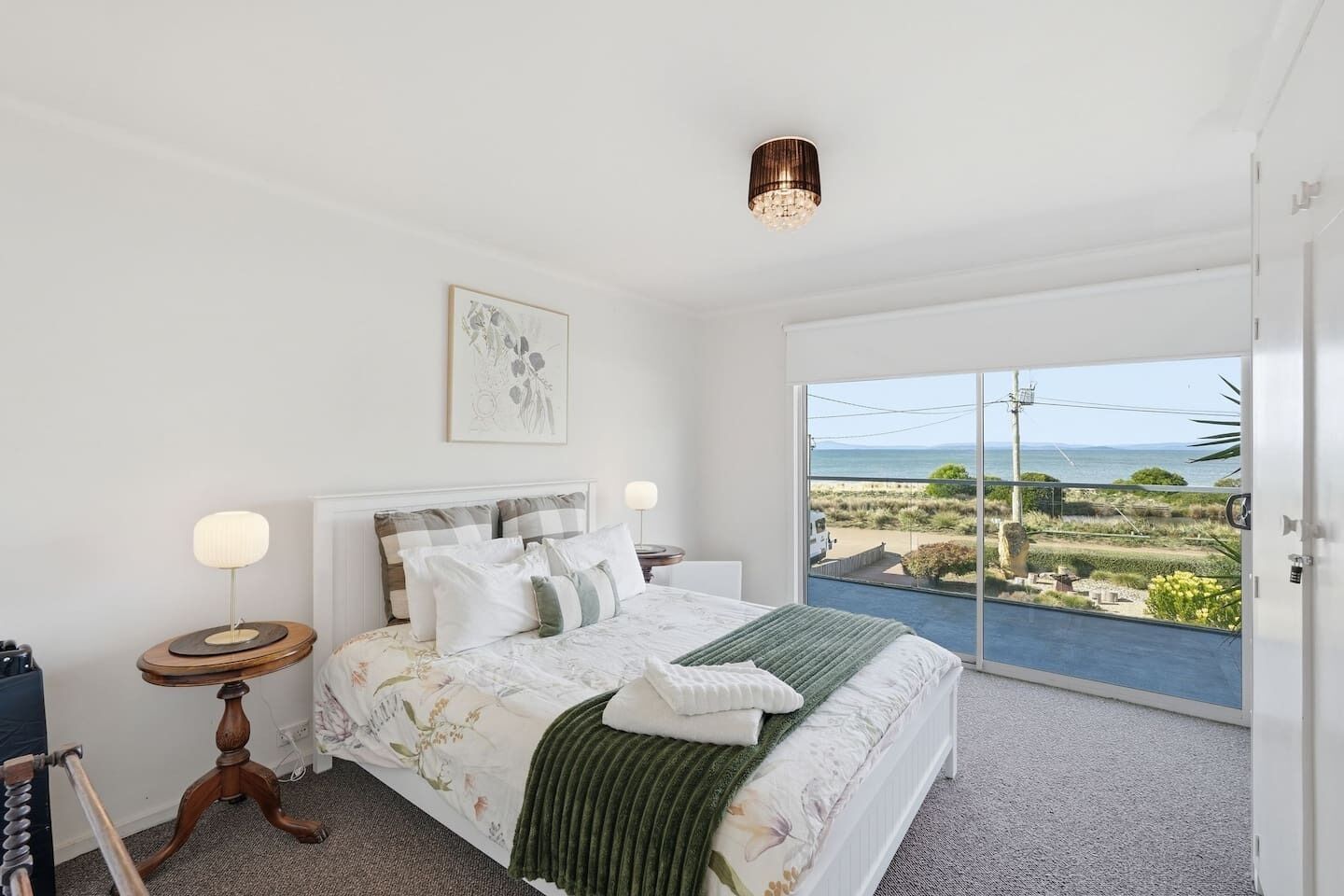 A bedroom with a bed, a bedside table, a lamp, a framed picture, and a view of the outdoors.