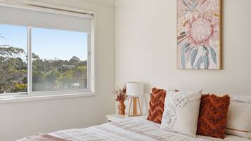 A bedroom with a large window, a bed with patterned bedding, a bedside table with a lamp, and a floral artwork on the wall.