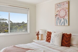 Ein Schlafzimmer mit großem Fenster, einem Bett mit gemusterten Betttextilien, einem Nachttisch mit Lampe und einem floralen Bild an der Wand.