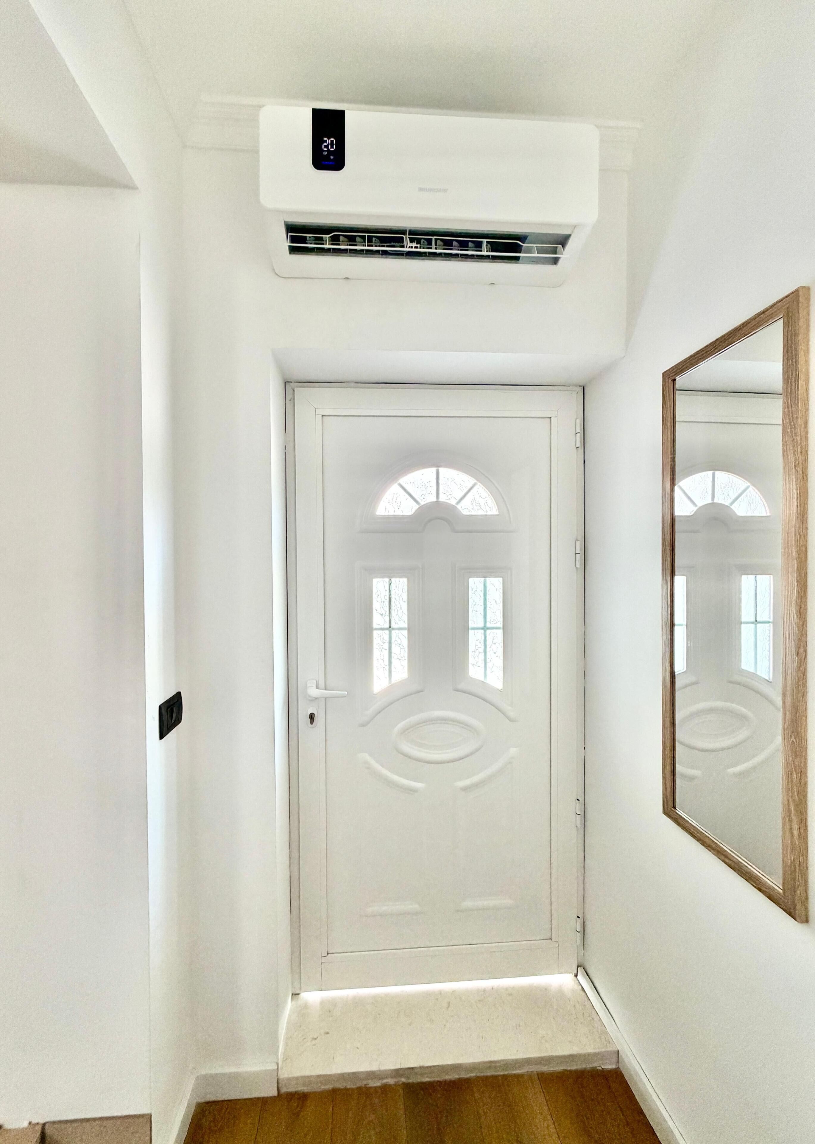 A white door with a small window, a wall-mounted air conditioning unit, and a mirror reflecting part of the room.