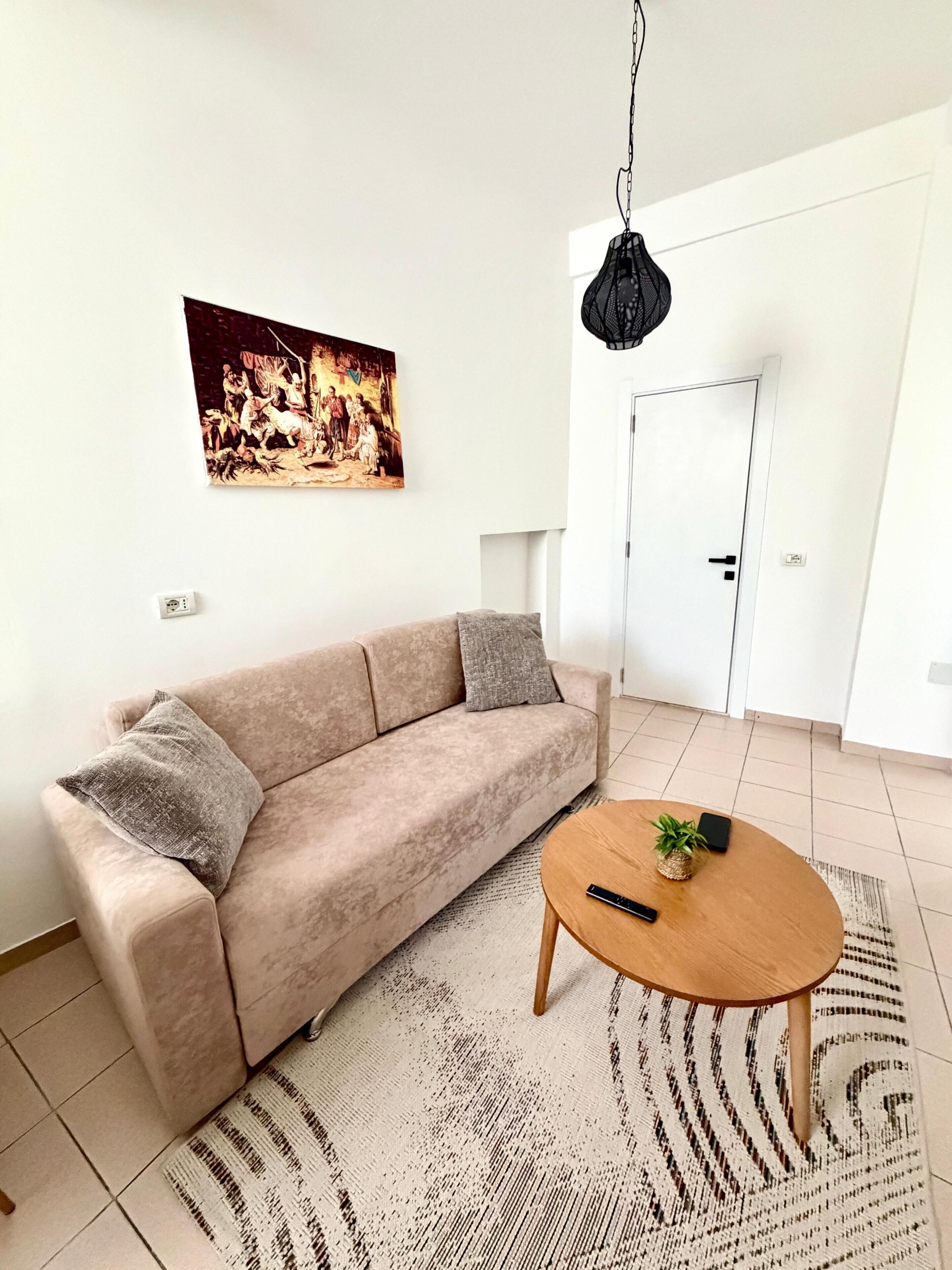 Un soggiorno con un divano beige, un tavolino rotondo in legno, una lampada a sospensione e un quadro appeso al muro.