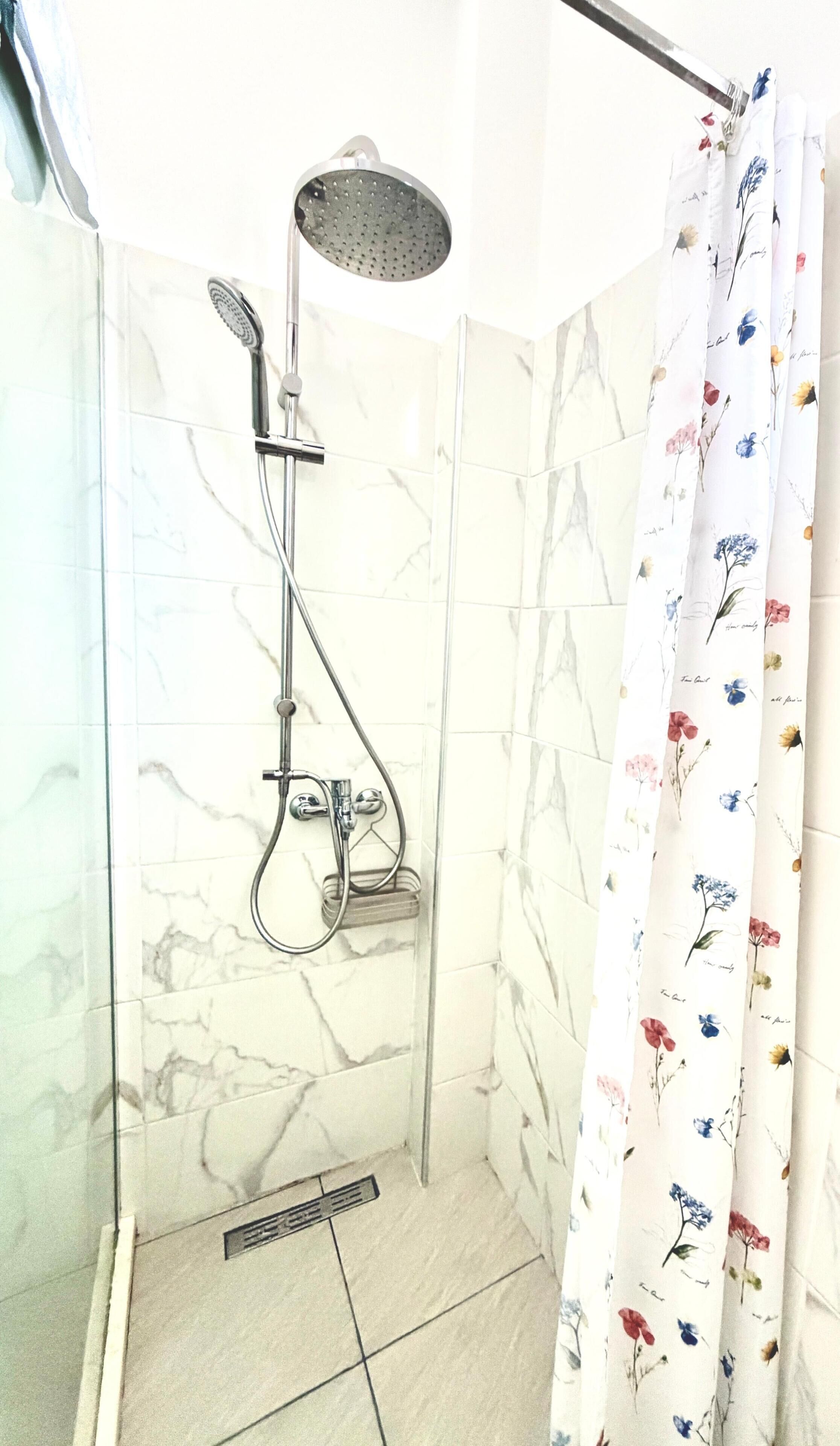 Eine Dusche mit Handbrause und Wandarmatur, ein Duschvorhang mit floralem Muster und ein gefliester Boden mit Marmoroptik.