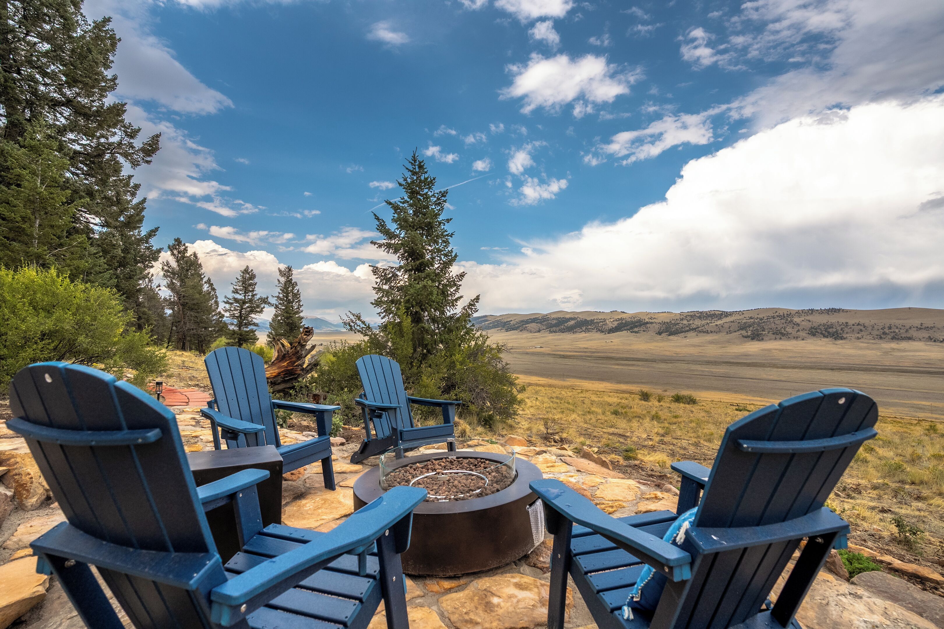 A seating area with Adirondack chairs around a fire pit, overlooking a vast landscape with hills and trees.