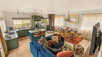 A modern living room with a blue sofa, a dining area with yellow chairs, a kitchen with green cabinets, and a stone fireplace.