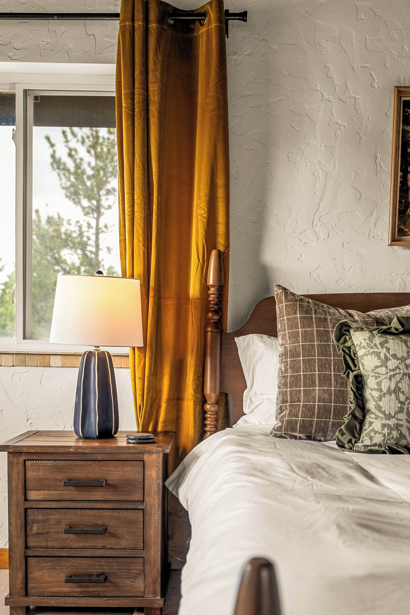 Een slaapkamer met een houten nachtkastje, een lamp, een bed met een witte dekbedovertrek, een geruit kussen en een raam met gordijnen.