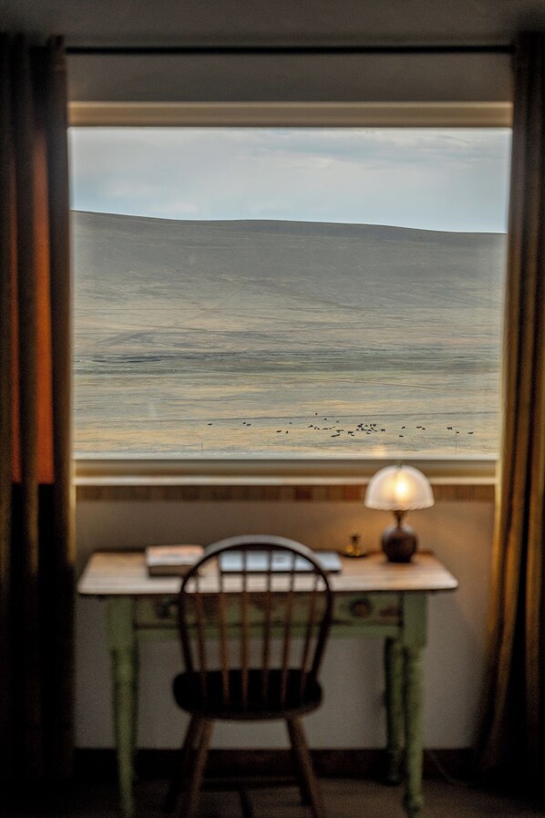 Une pièce avec vue sur un paysage plat, visible par une fenêtre, comprenant une chaise et une table avec une lampe.