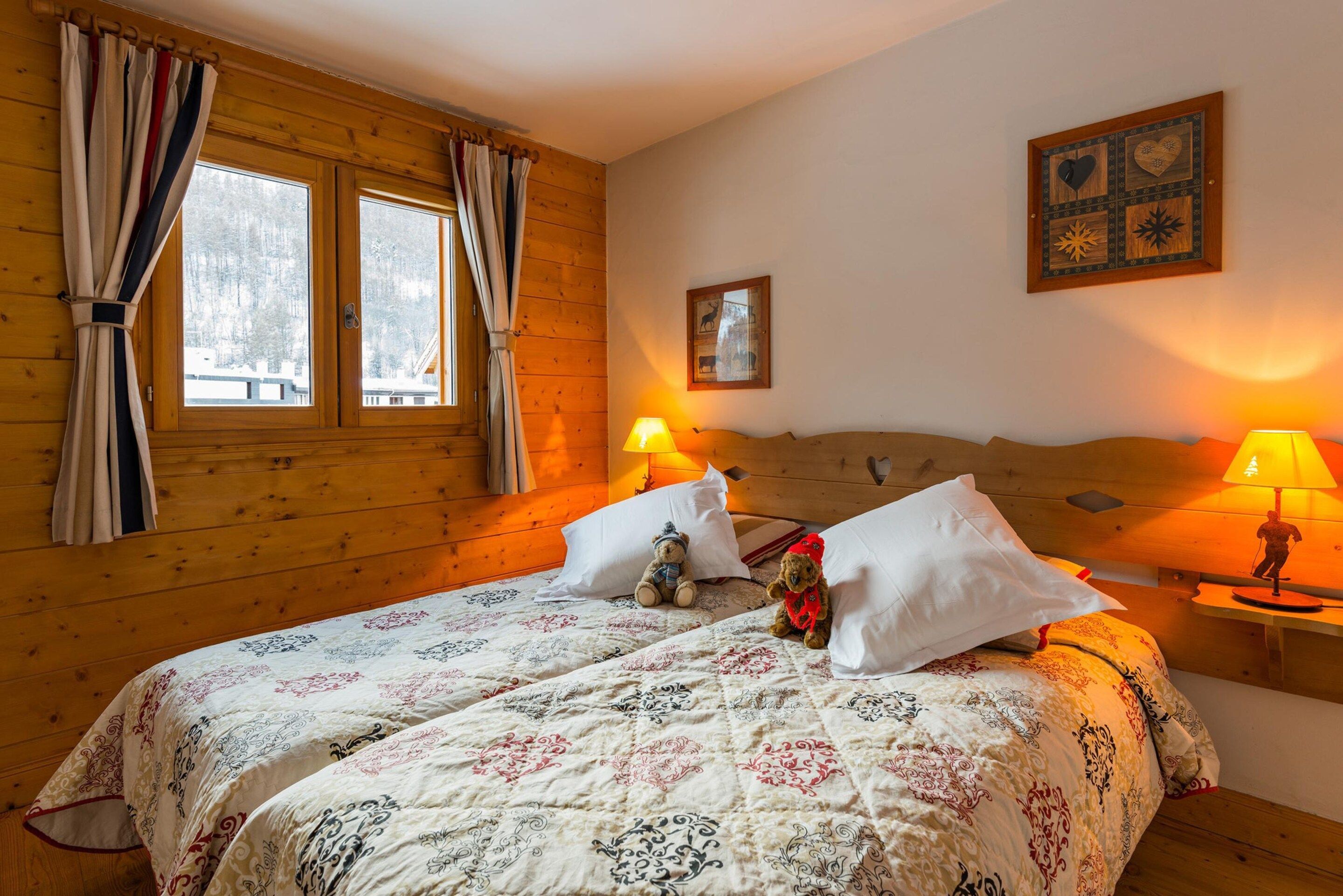 A cozy wooden room with a bed, two bedside lamps, a window with curtains, and a wall with a decorative panel.