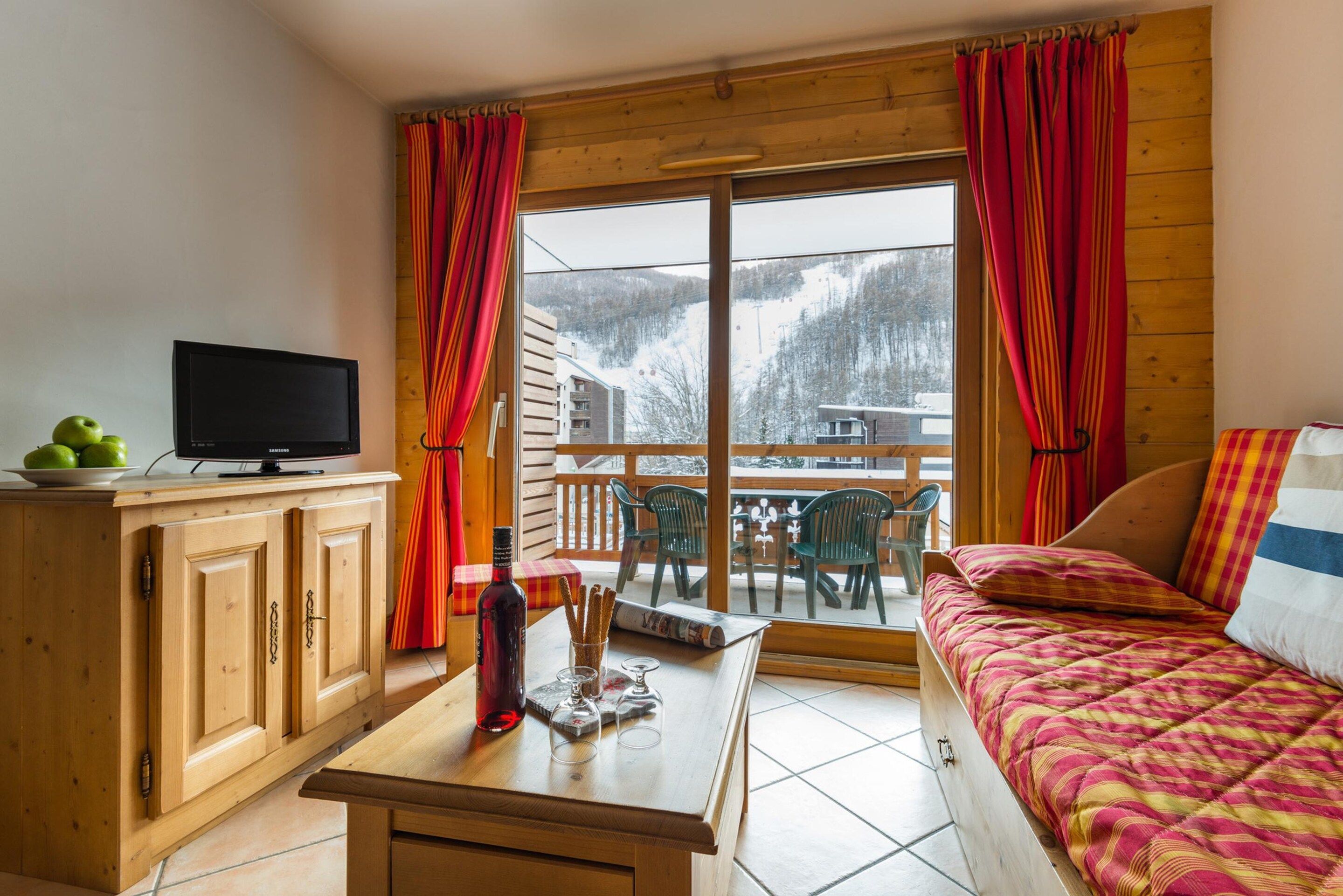 A cozy room with a bed, a television on a wooden cabinet, a table with a bottle and glasses, and a view of a snowy landscape through large windows.