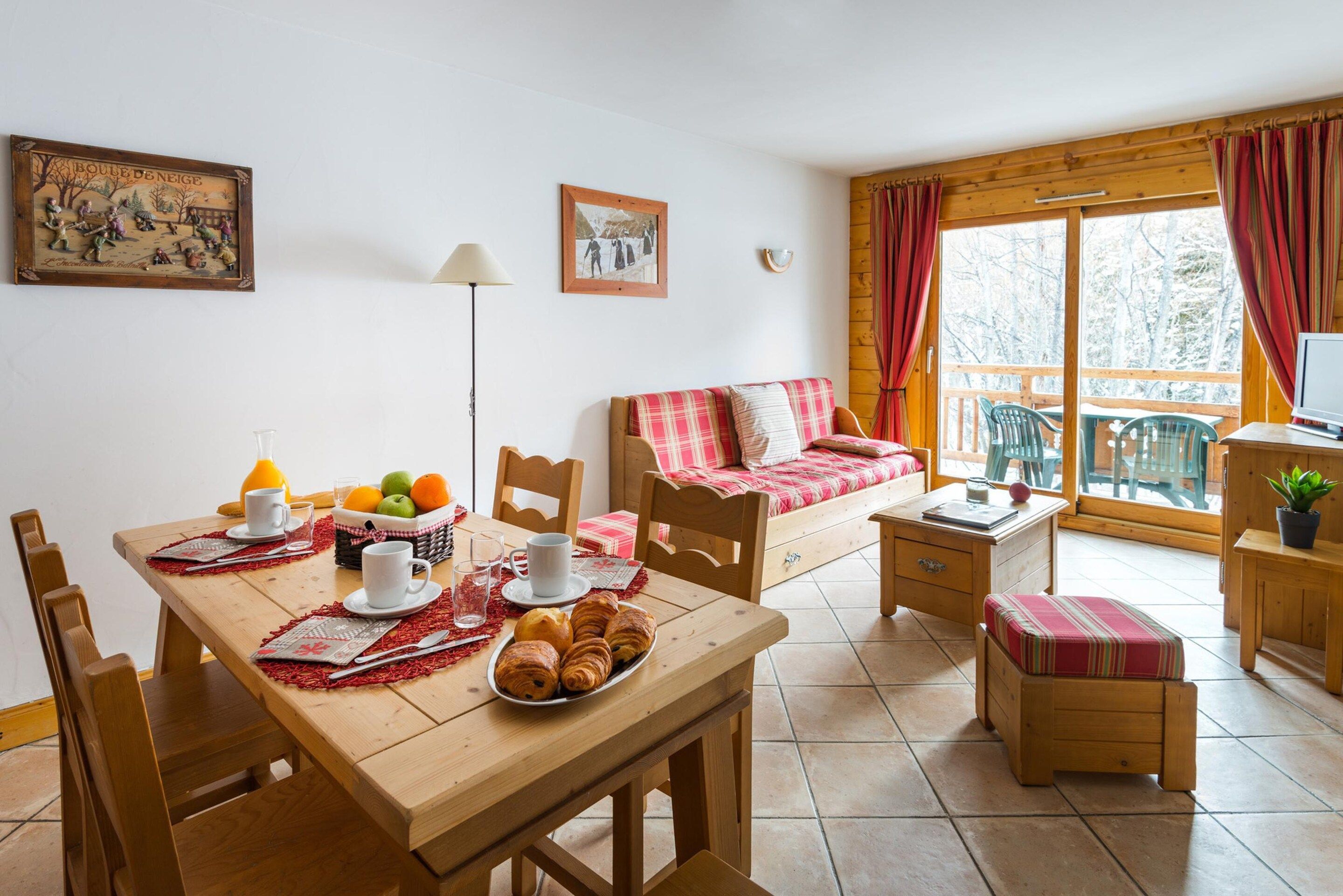 A cozy room with a wooden dining table set for breakfast, a sofa with a plaid blanket, and a window with curtains.