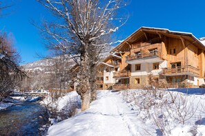 A wooden chalet with multiple balconies, a stone foundation, and a view of a snow-covered landscape with a river.