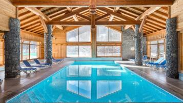 An indoor swimming pool with clear blue water, wooden beams, stone pillars, and blue lounge chairs.