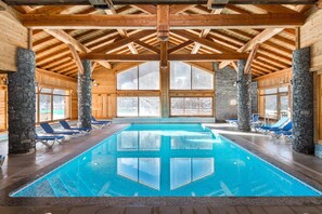 An indoor swimming pool with clear blue water, wooden beams, stone pillars, and blue lounge chairs.