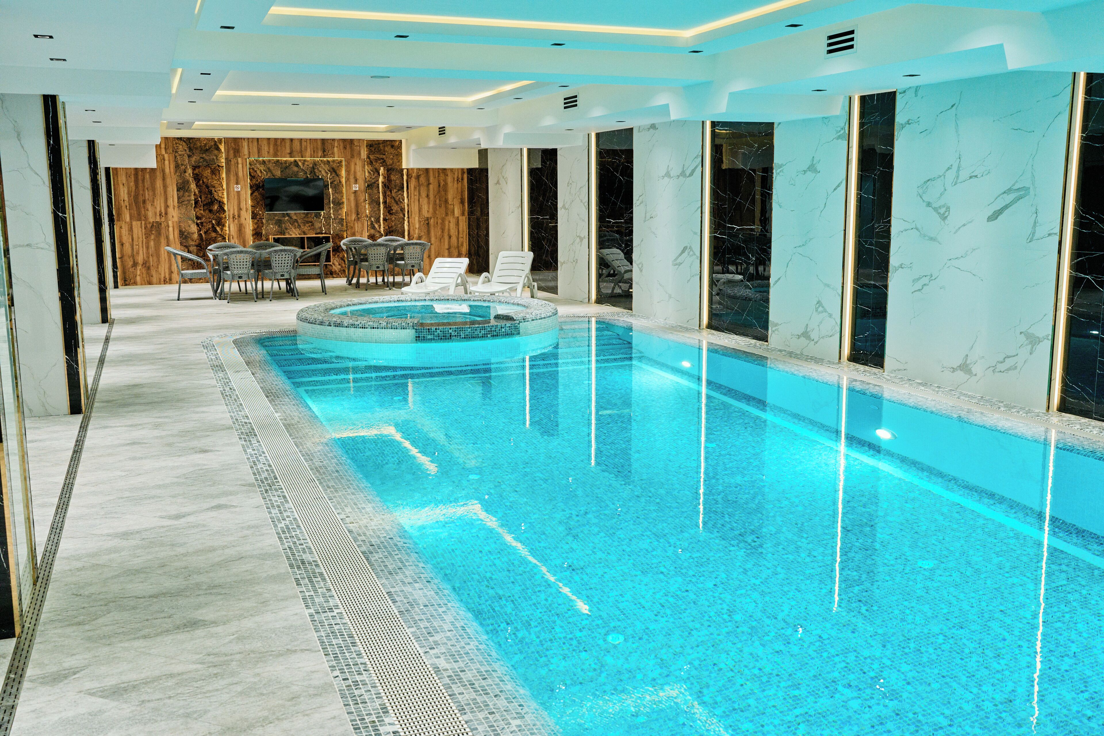 A modern indoor swimming pool with clear water, surrounded by lounge chairs and a wooden wall with a television.