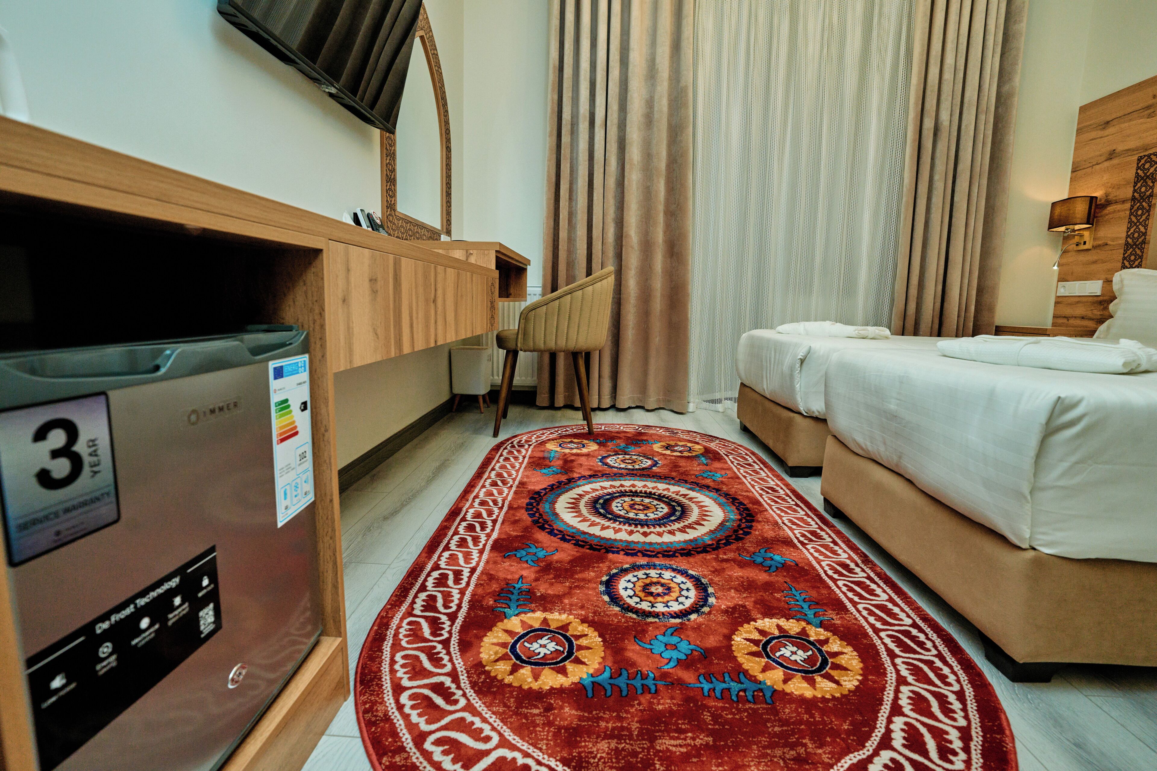 A hotel room with a bed, a desk with a computer, a TV mounted on the wall, and a patterned rug.