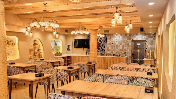 A restaurant interior with wooden tables and patterned chairs, a bar area, and a television mounted on the wall.