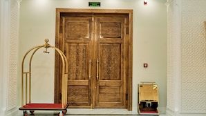 A hotel entrance with a double wooden door, a gold-colored luggage cart, and a vacuum cleaner.