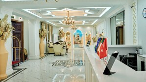 A hotel lobby with a reception desk, chandeliers, decorative plants, and a view of the lobby through an archway.