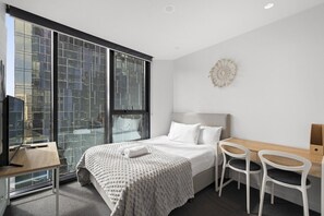 Una habitación de hotel moderna con un ventanal grande, una cama con ropa de cama blanca, un escritorio con una silla y una mesita.