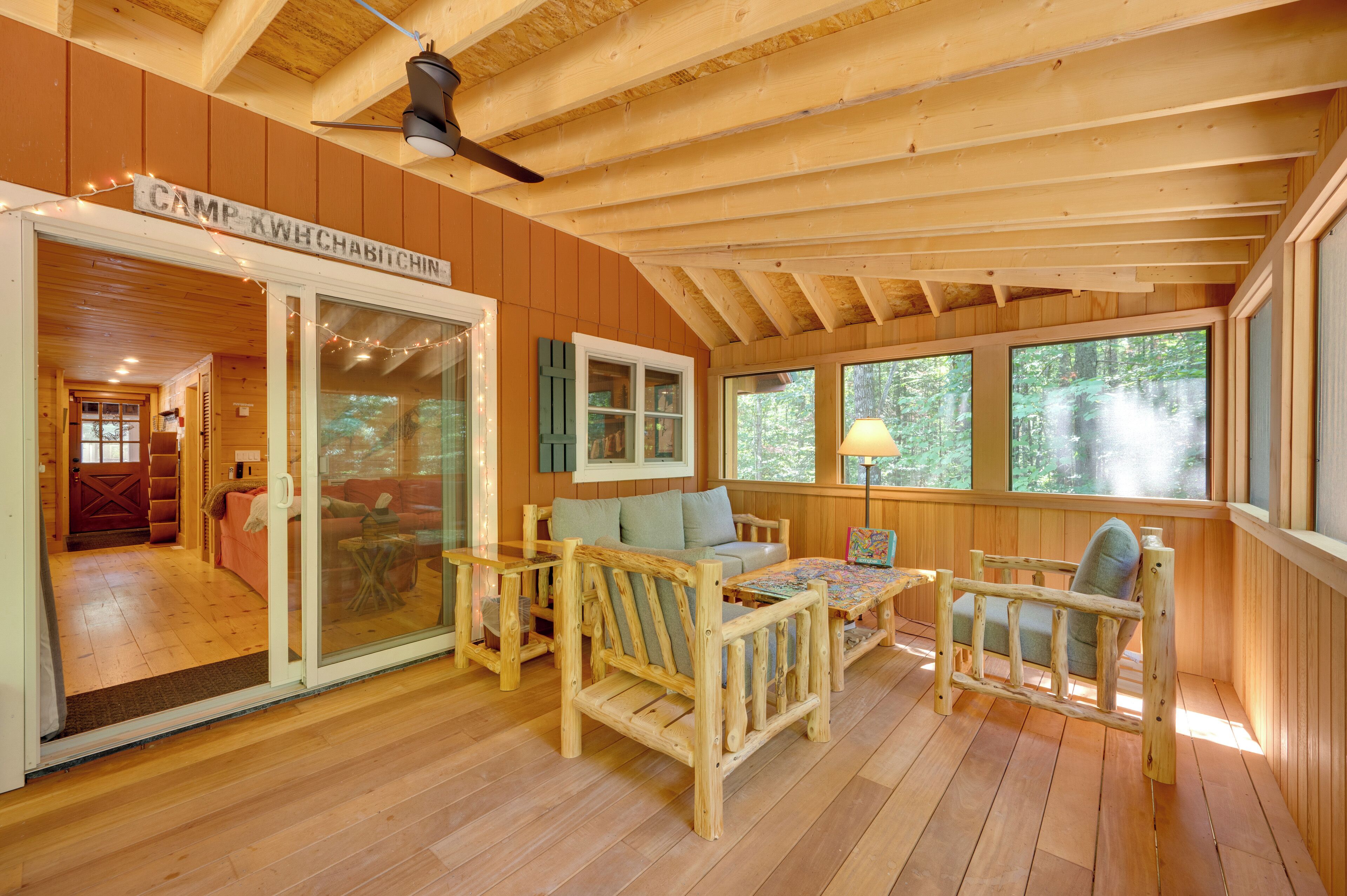 A cozy cabin interior with a wooden ceiling, a ceiling fan, a sofa, two chairs, a puzzle table, and a large window offering a view of the forest.