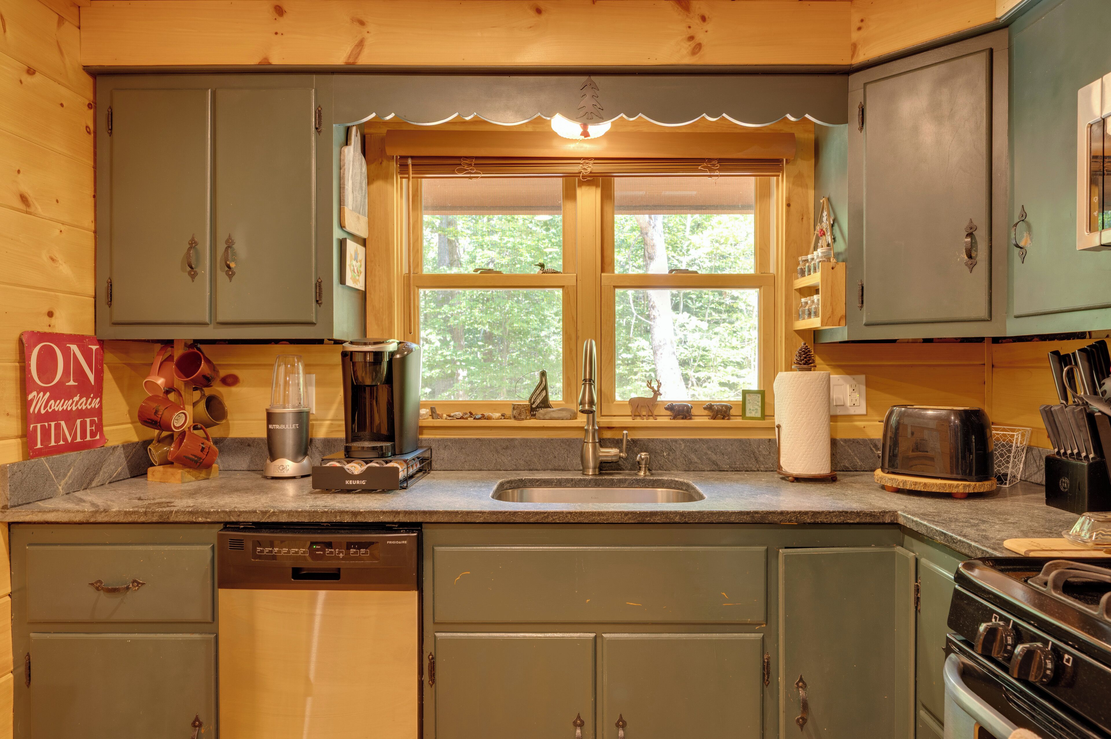 Una cocina con armarios de madera, lavavajillas integrado, cafetera y ventana con vista a árboles.