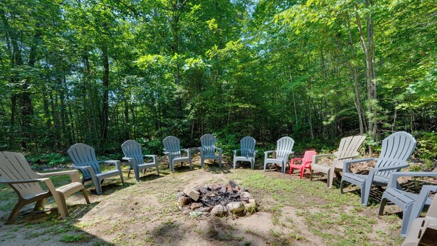 A group of Adirondack chairs arranged around a campfire in a forested area. - North Conway: Updated 3BR Cabin Sleeps 9, Fire Pit (North Conway)