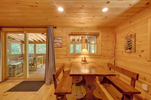 L’intérieur d’une cabane avec une table à manger en bois, des bancs, une lampe et une fenêtre donnant sur la forêt.