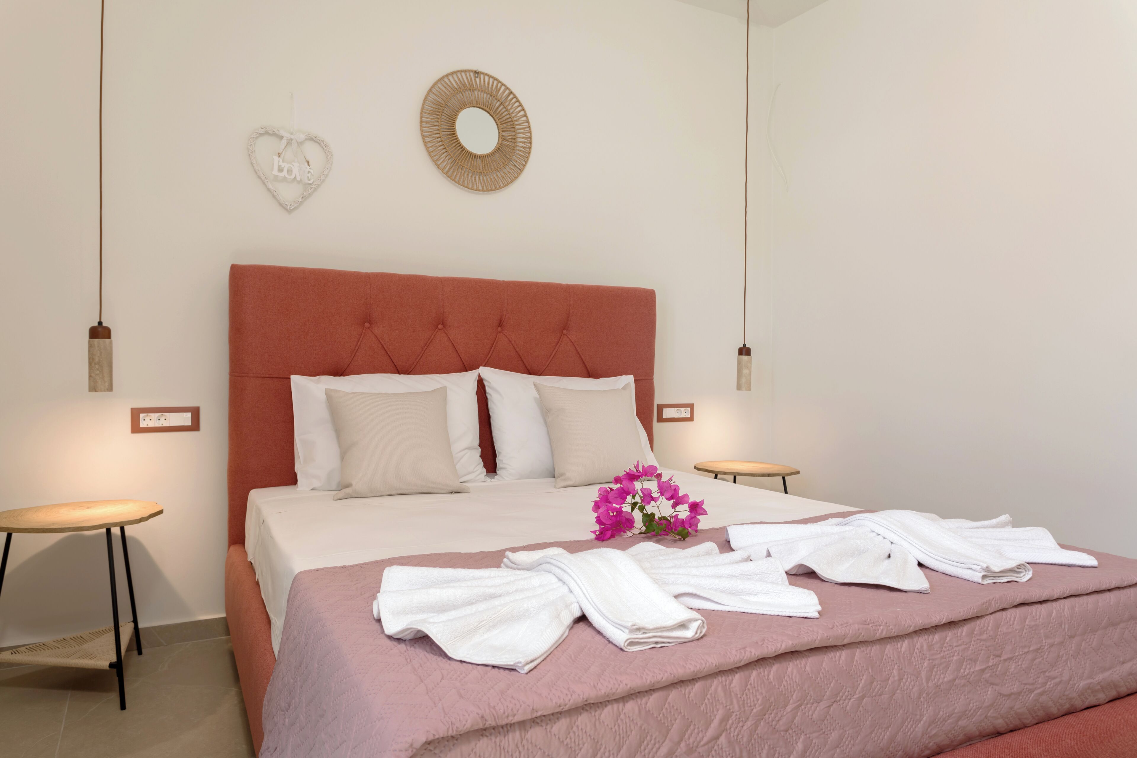 A neatly made bed with white linens, a pink blanket, and a bouquet of pink flowers.