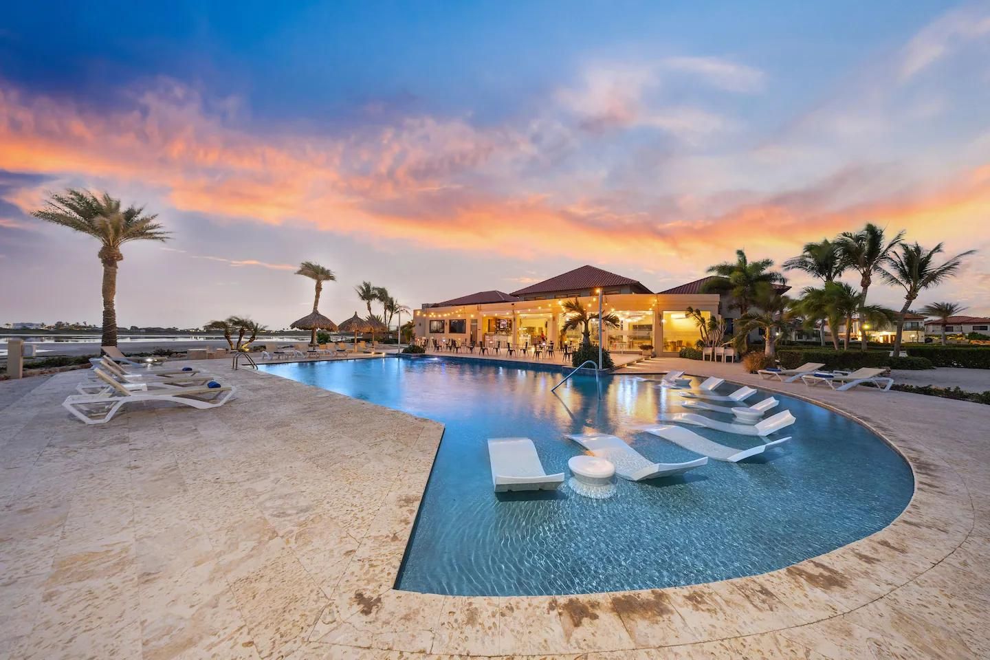 A resort pool with lounge chairs, palm trees, and a building with a red roof.