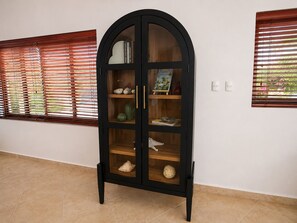 A black armoire with glass doors, placed in a hallway with tiled flooring and windows with blinds. - Stylish 3BR villa in Gold Coast Aruba with private pool, gym, tennis courts and gated resort amenities. (Noord)