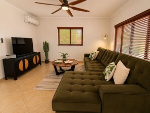 A living room with a sectional sofa, a coffee table, a television on a black cabinet, a ceiling fan, and a window with blinds. - Stylish 3BR villa in Gold Coast Aruba with private pool, gym, tennis courts and gated resort amenities. (Noord)