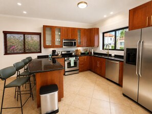 A modern kitchen with wooden cabinets, stainless steel appliances, a central island with bar stools, and a view of the outside through a window. - Stylish 3BR villa in Gold Coast Aruba with private pool, gym, tennis courts and gated resort amenities. (Noord)