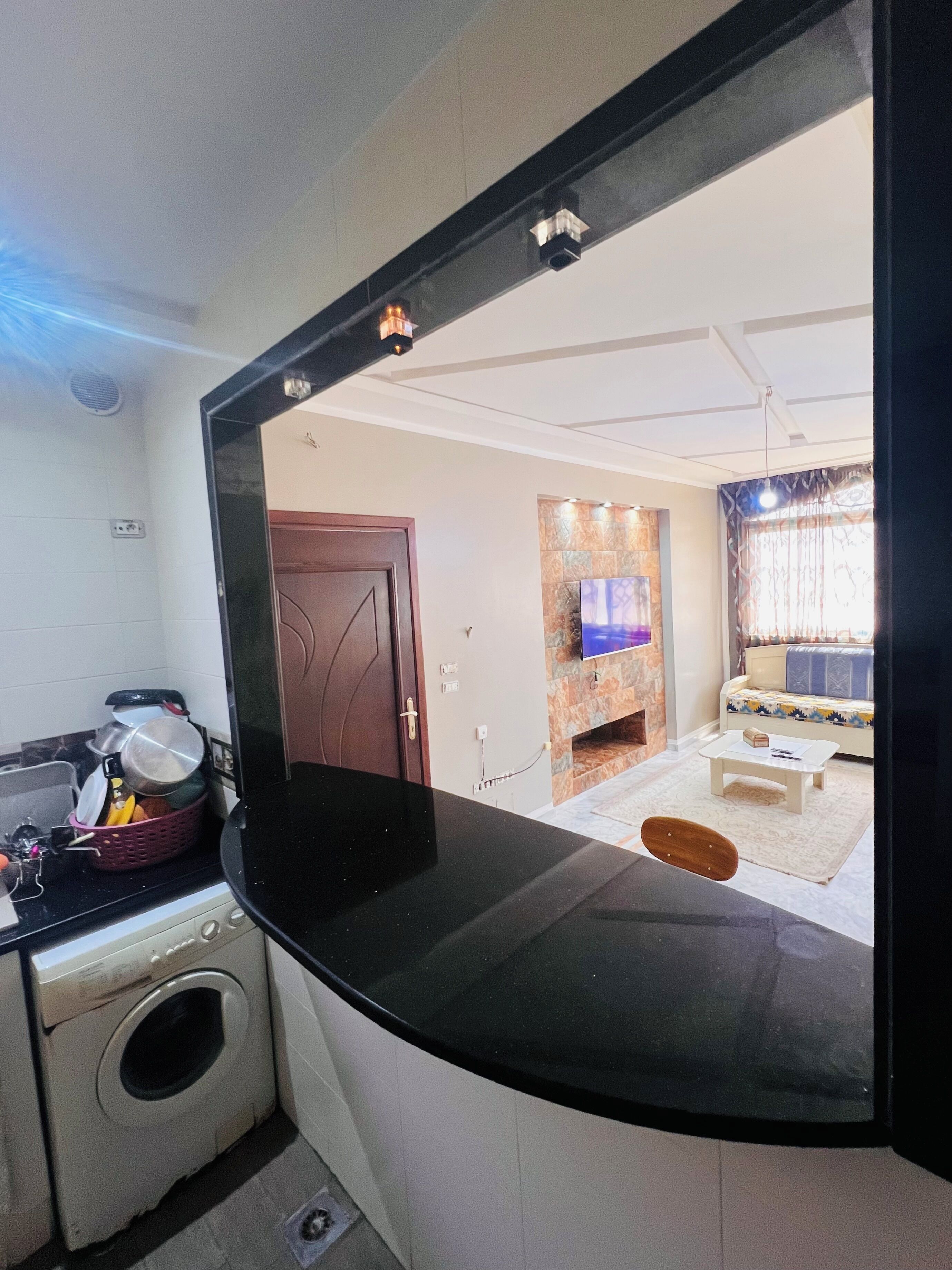 Eine Küche mit Waschmaschine, ein großer Spiegel, der ein Wohnzimmer mit Sofa und Fernseher reflektiert.