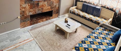 A living room with a stone fireplace, a flat-screen TV mounted on the wall, a sofa with patterned cushions, and a coffee table with a remote control.