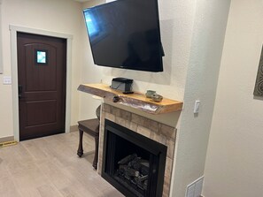 A living room with a flat-screen TV mounted above a fireplace, a wooden mantel, and a brown door.