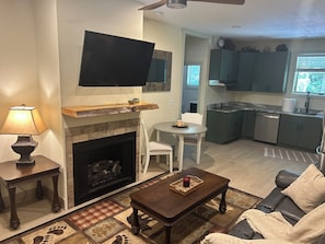 Uma sala de estar com lareira, televisão montada acima, mesa de jantar com cadeiras e cozinha com armários e janela.
