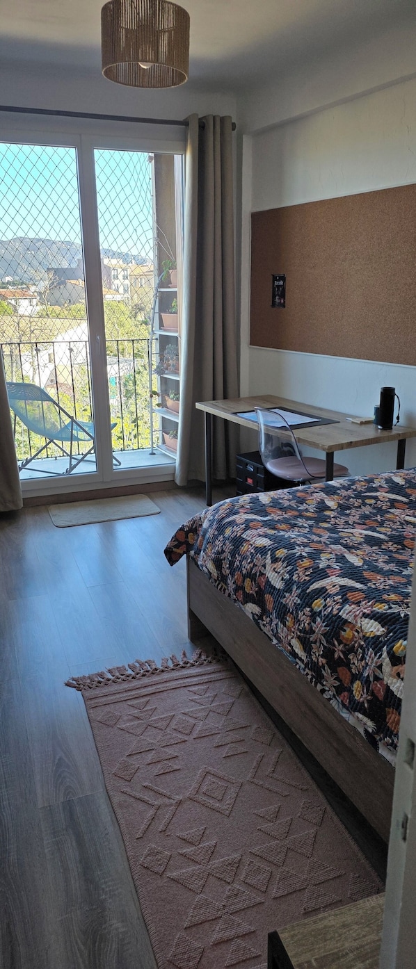 A bedroom with a bed, a desk, a chair, a hanging light fixture, and a view of the outside. - Bed and Breakfast for Solo Female Travelers (Marseille)