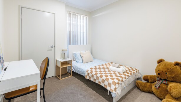 Een slaapkamer met een bed, een bureau, een stoel, een teddybeer en een raam met jaloezieën.