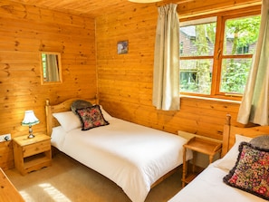 Ein Schlafzimmer einer Holzhütte mit zwei Betten, einem Nachttisch, einem Beistelltisch, einem Fenster mit Vorhängen und einem Spiegel.